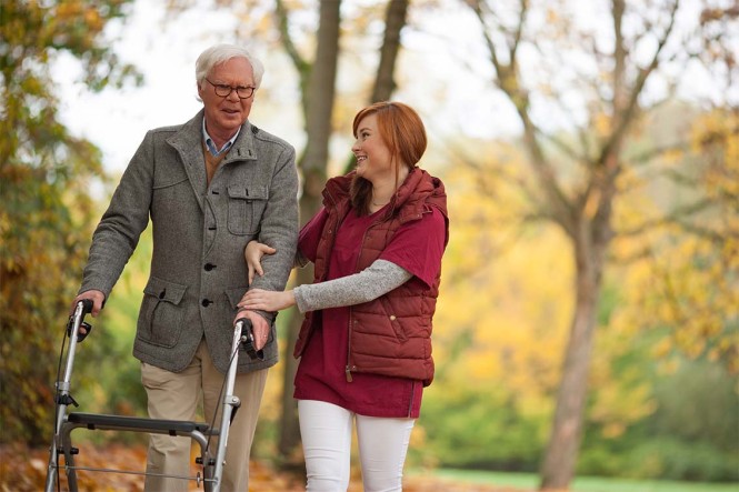 Ein älterer Mann mit Rollator und eine Pflegerin gehen spazieren.