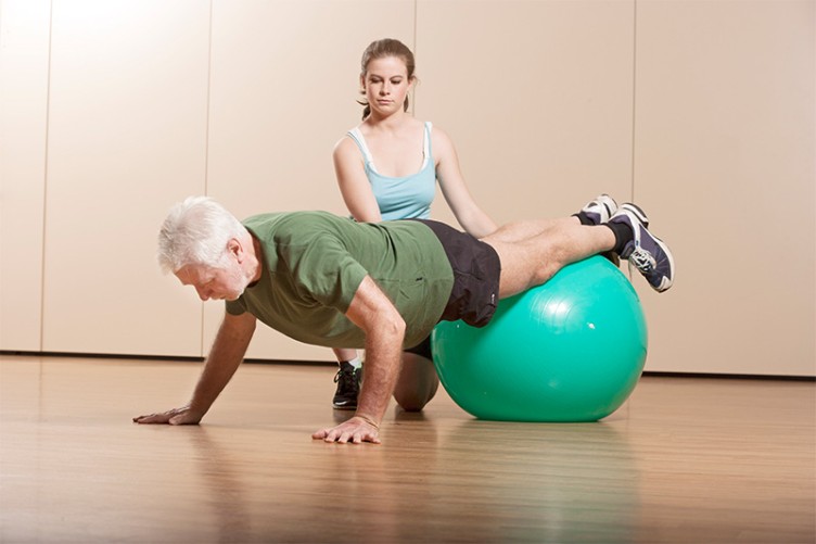 Eine Trainerin übt mit einem älteren Mann und einem Medizinball